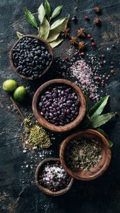 A wooden bowl filled with black beans surrounded by fresh onions, tomatoes, and herbs on a rustic wooden table.